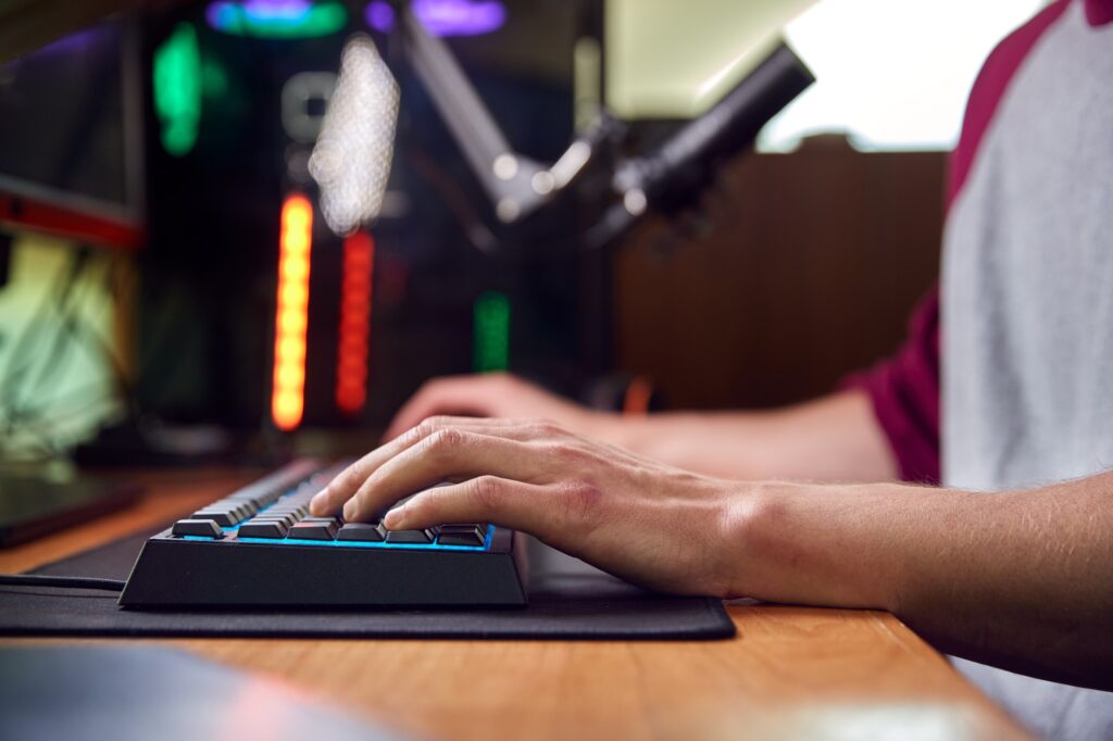 Close Up Of Hands On Keyboard As Man Games At Home Sitting At Desk With Multiple Monitors